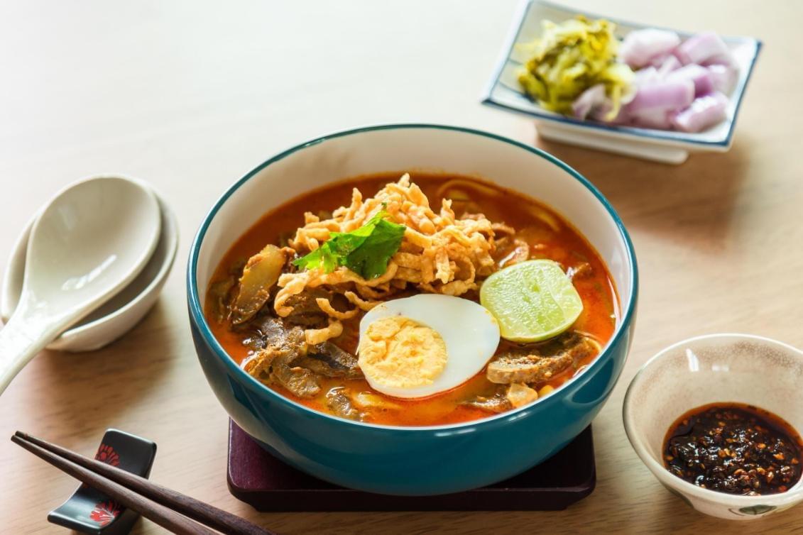  This overhead image showcases a vibrant bowl of Khao Soi, a Northern Thai curry noodle soup, presented in a teal-rimmed bowl. The soup contains egg noodles in a rich, reddish-orange curry broth, topped with meat, a wedge of lime, half of a hard-boiled egg.