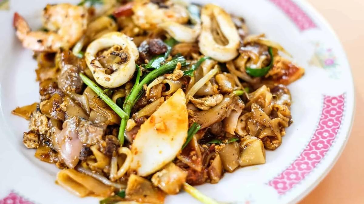 This image depicts a plate of Char Kway Teow, a popular Southeast Asian stir-fried noodle dish. The dish features flat rice noodles wok-fried with dark soy sauce and is topped with ingredients such as prawns, squid rings, and green vegetables.