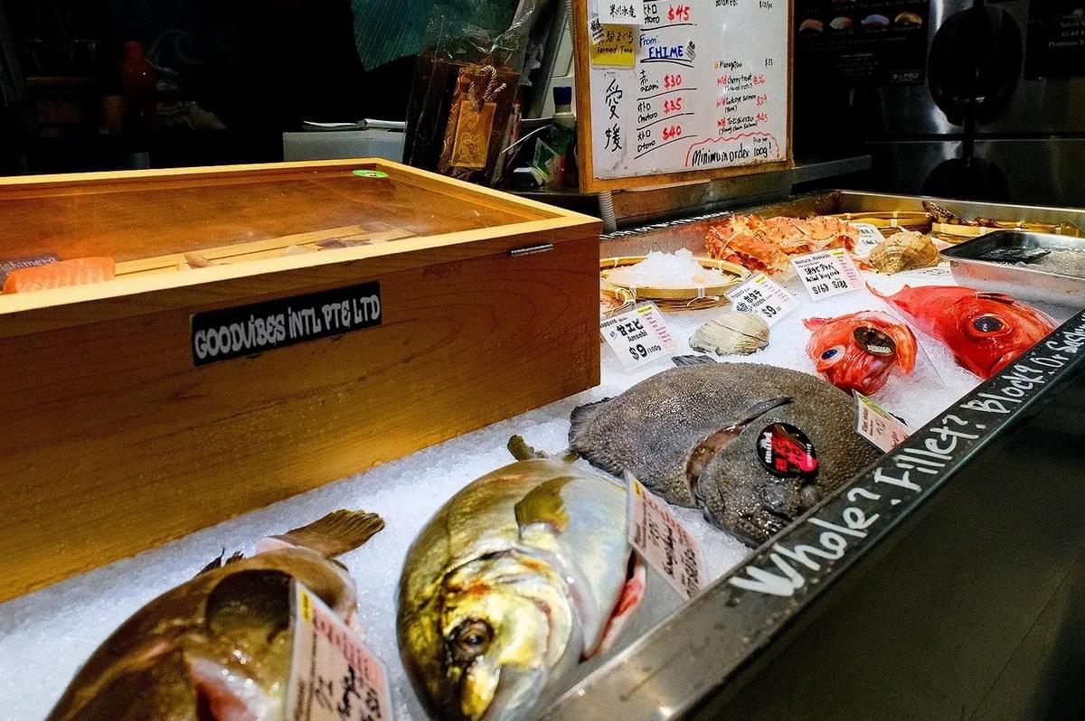 A seafood counter displays a variety of fresh fish, crab, and shellfish laid out on crushed ice. Small price signs sit alongside the catches, with handwritten menu boards visible in the background.