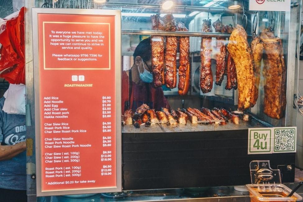 The image showcases a "Roast Paradise" stall featuring rows of glistening roasted meats suspended behind a glass display window. A detailed red menu board lists various rice and noodle options, with prices ranging from $4.50 to $18.00 depending on the portion size.