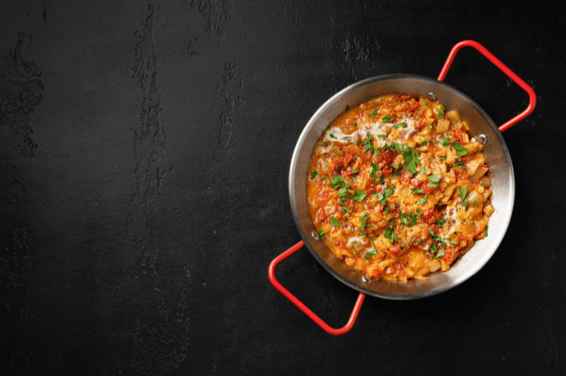A pan of Turkish menemen—scrambled eggs simmered with tomatoes, onions, and peppers in a creamy, herb-topped stew—rests on a dark surface, its red handles echoing the dish’s vibrant warmth.