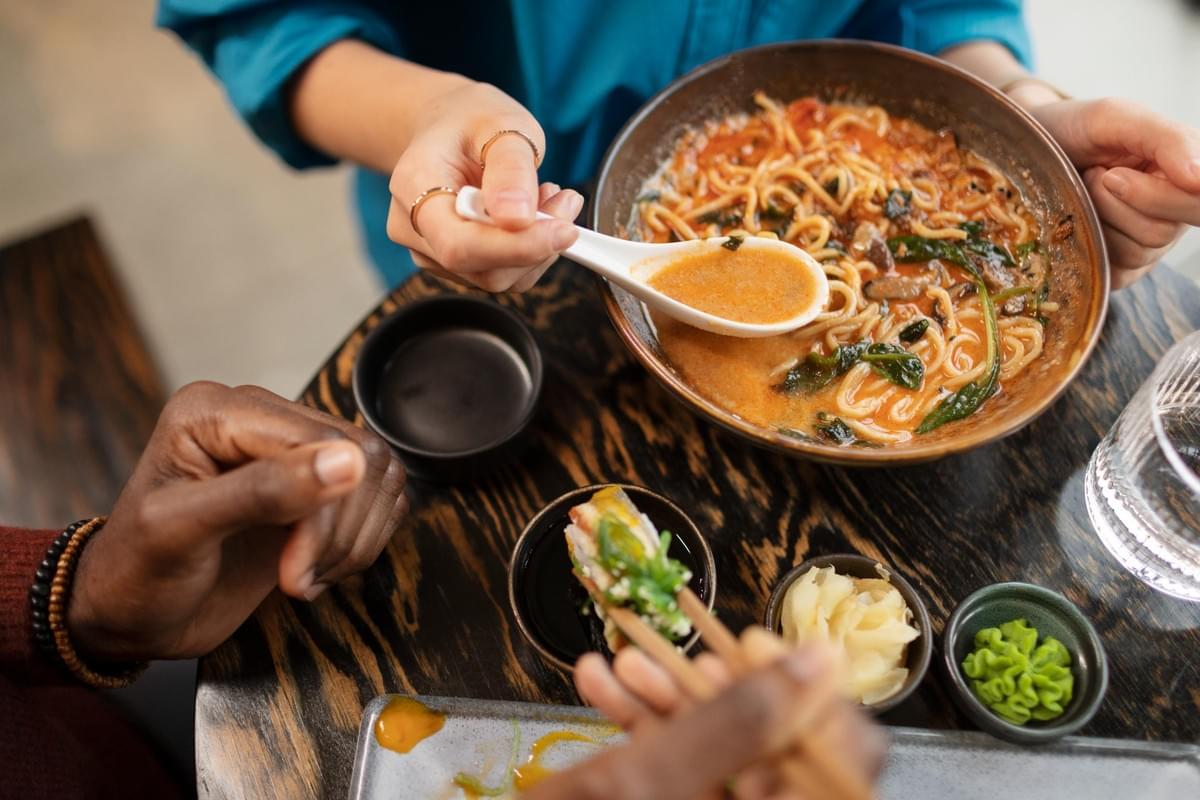 The image features a close-up, overhead view of two people sharing a meal at a dark wooden table. One person uses a ceramic spoon to scoop broth from a large bowl of noodle soup, while the other uses chopsticks to dip a piece of sushi into sauce.