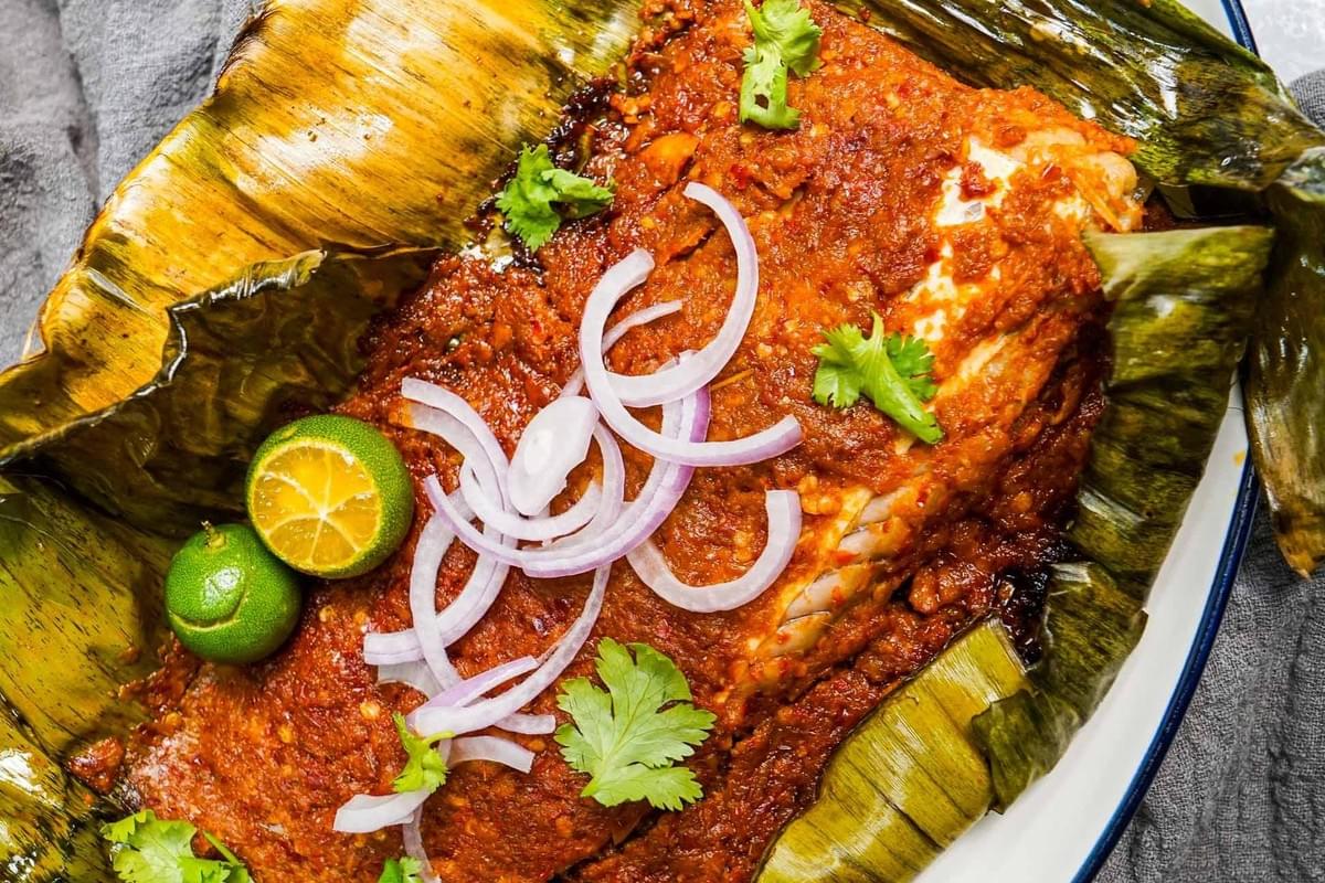 This image features a piece of grilled stingray smothered in a vibrant, spicy sambal sauce and served on a charred banana leaf. It is garnished with fresh red onion rings, sprigs of cilantro, and halved calamansi limes for a bright, acidic finish.