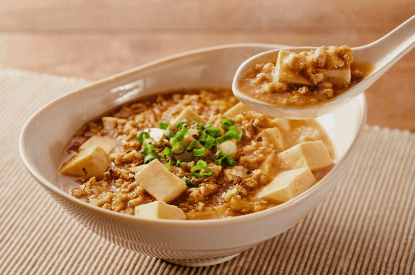 A white ceramic spoon lifting a portion of silky Mapo Tofu from a bowl, showing the cubes of soft tofu mixed with minced meat in a savory brown sauce topped with fresh green onions.