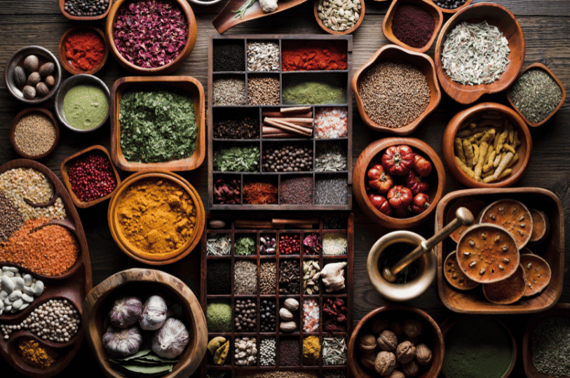 A high-angle, organized overhead shot of various spices in wooden bowls and a rustic apothecary-style wooden box. Ingredients include star anise, cinnamon sticks, garlic bulbs, dried flower petals, peppercorns, and diverse colorful powders against a dark wood background.
