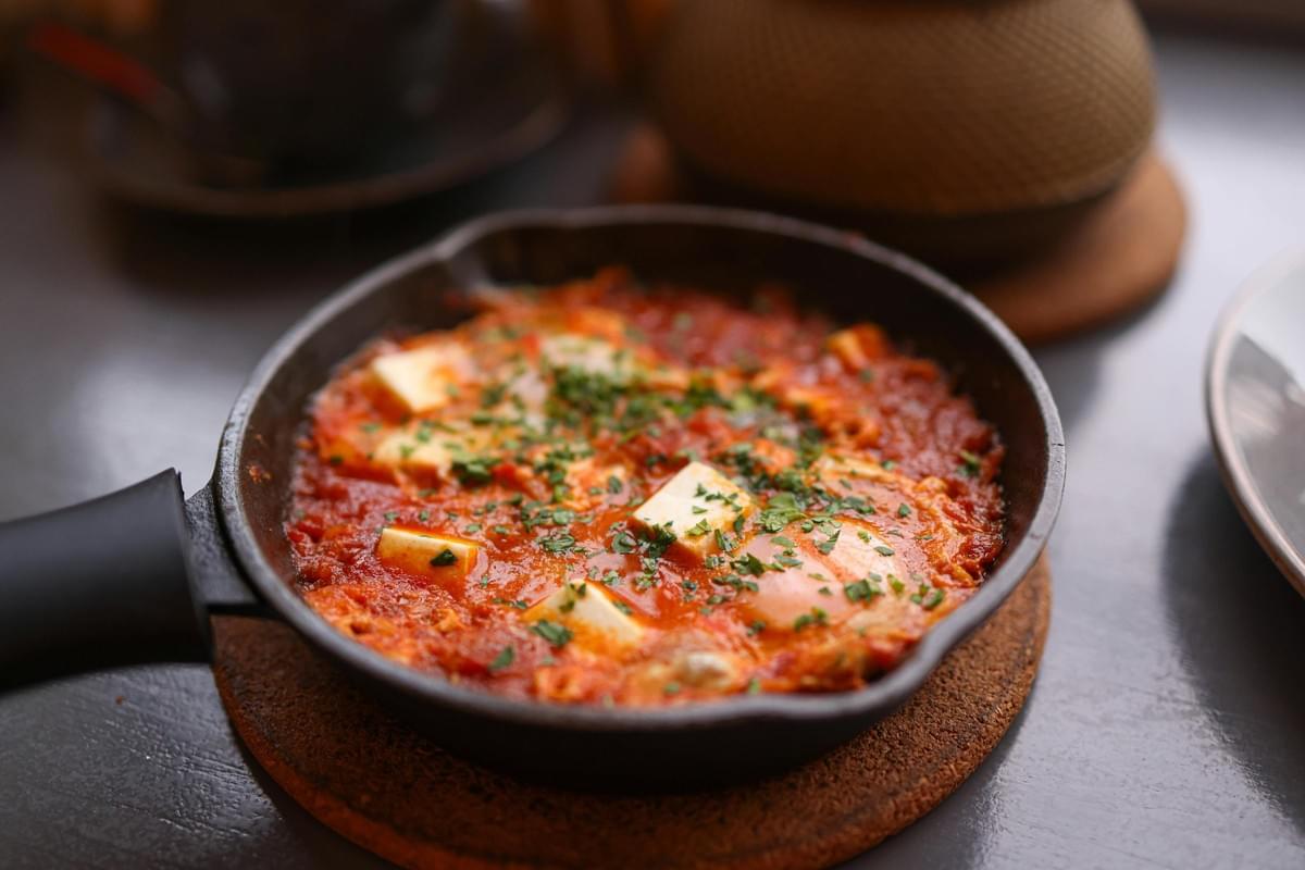 A small black skillet holds a warm, tomato-based dish featuring poached eggs and cubes of white cheese, generously garnished with chopped green herbs. The pan rests on a round cork coaster, set against a background that includes a textured teapot, suggesting a cozy meal setting.