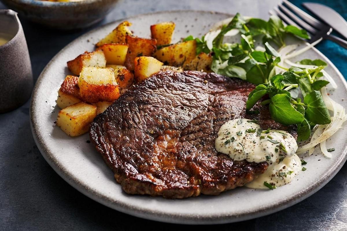 A perfectly seared rib-eye steak sits on a grey ceramic plate, topped with a dollop of herb butter. It is served alongside golden, crispy cubed potatoes and a fresh green salad with thinly sliced onions.