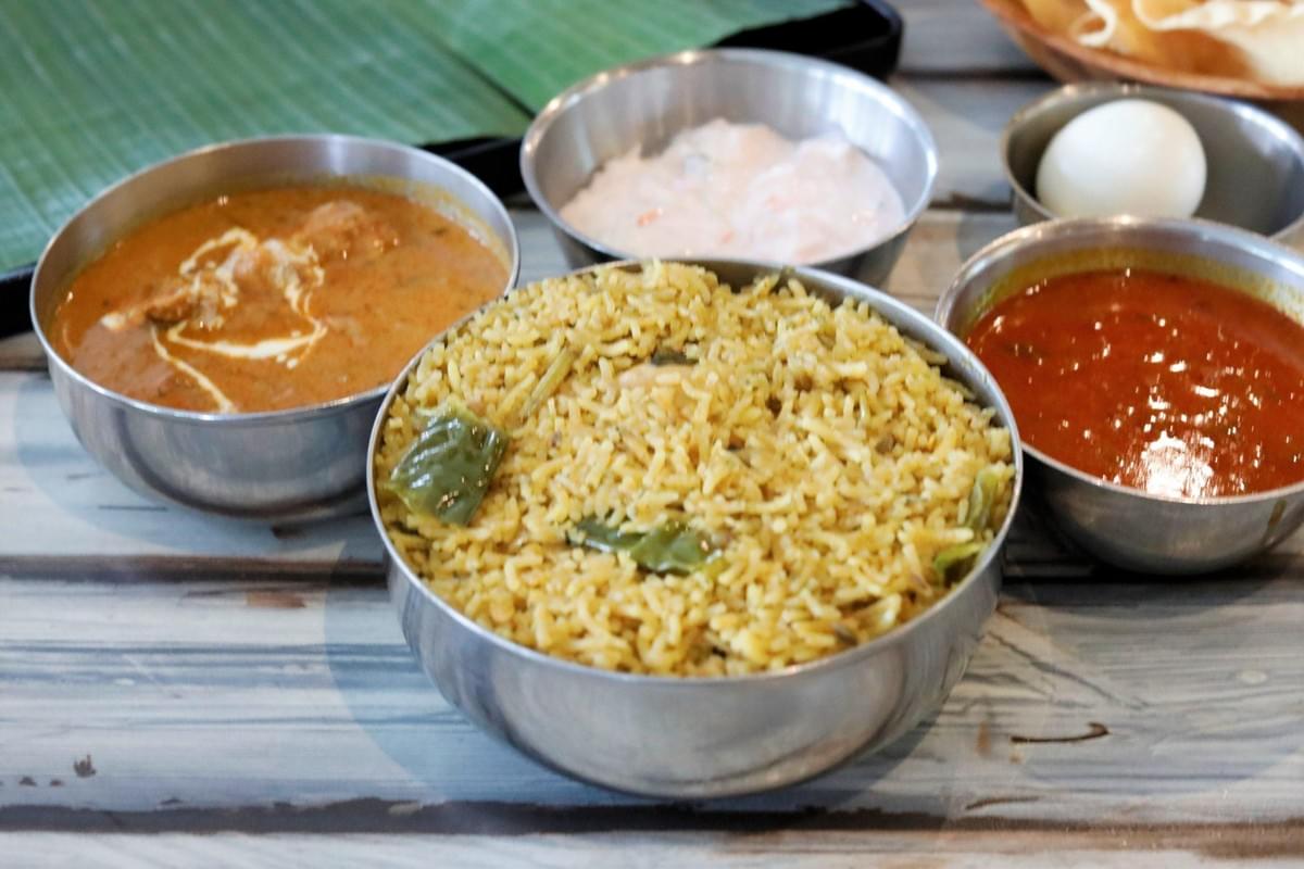 The image displays a South Indian meal served in multiple stainless steel bowls on a wooden table. A large bowl of seasoned biryani rice sits in the foreground, accompanied by bowls of creamy chicken curry, raita, a hard-boiled egg, and a spicy red gravy.
