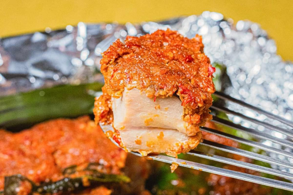 A fork holds up a tender piece of grilled stingray topped with a thick, vibrant red layer of spicy sambal. The fish is served on a banana leaf over aluminum foil, showcasing its flaky white texture beneath the aromatic chili paste.