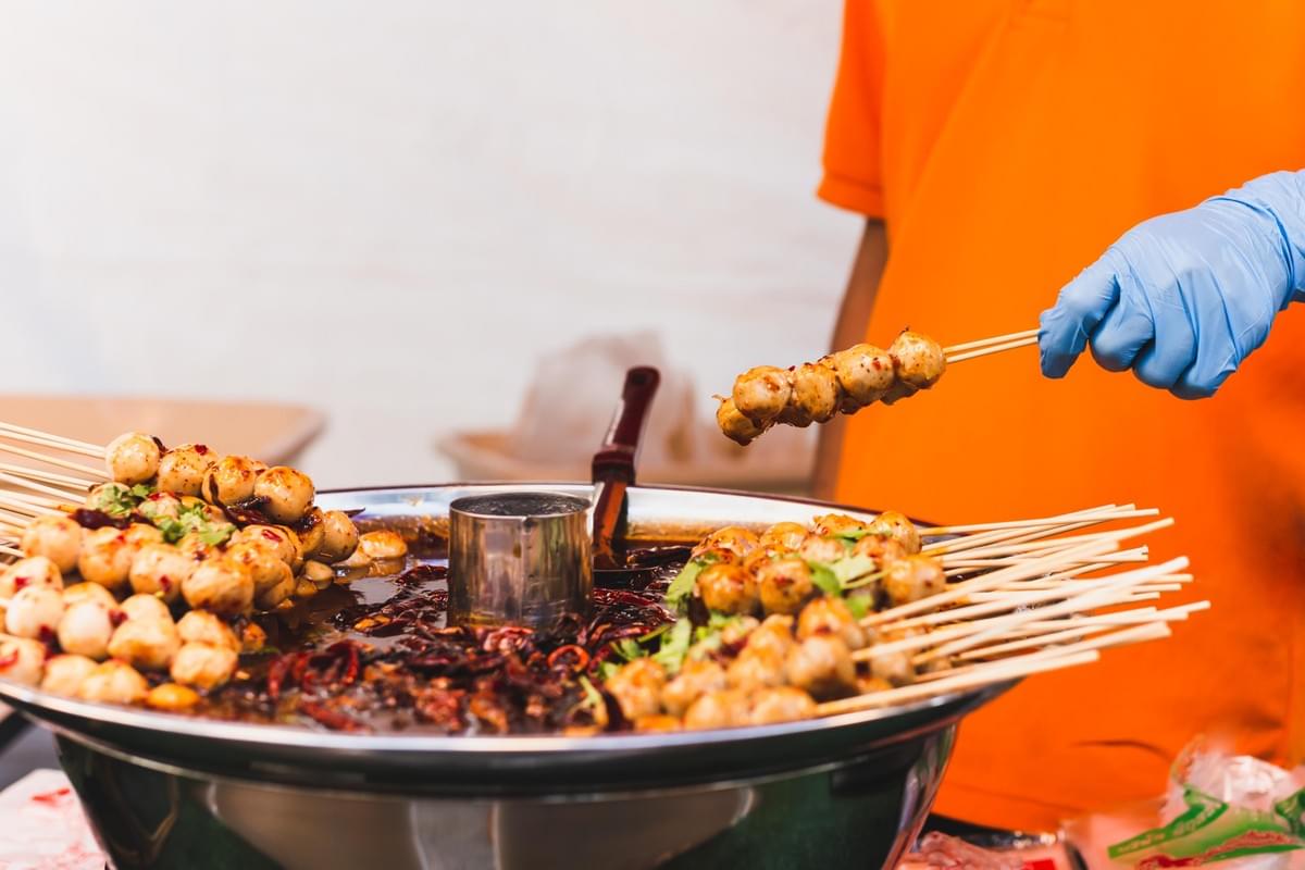 A vendor wearing a blue latex glove holds up skewers of meatballs over a large metal basin filled with a dark, spicy sauce containing visible dried chilies. Numerous other skewers rest along the rim of the bowl, soaking in the rich glaze and garnished with fresh green herbs, characteristic of a vibrant street food stall.