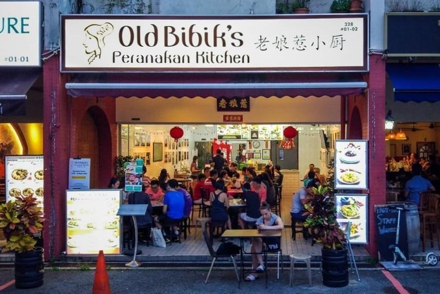 The storefront of Old Bibik’s Peranakan Kitchen features a large, illuminated sign above an open dining area filled with patrons enjoying their meals. Red lanterns hang from the ceiling, while outdoor menu displays and potted plants frame the entrance to the lively restaurant.