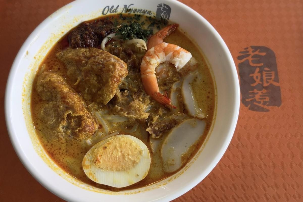 A steaming bowl of laksa from Old Nyonya features a rich, orange coconut curry broth topped with a halved hard-boiled egg, prawns, and tau pok. The dish is presented on an orange textured table, accompanied by a dollop of sambal and finely chopped laksa leaves.