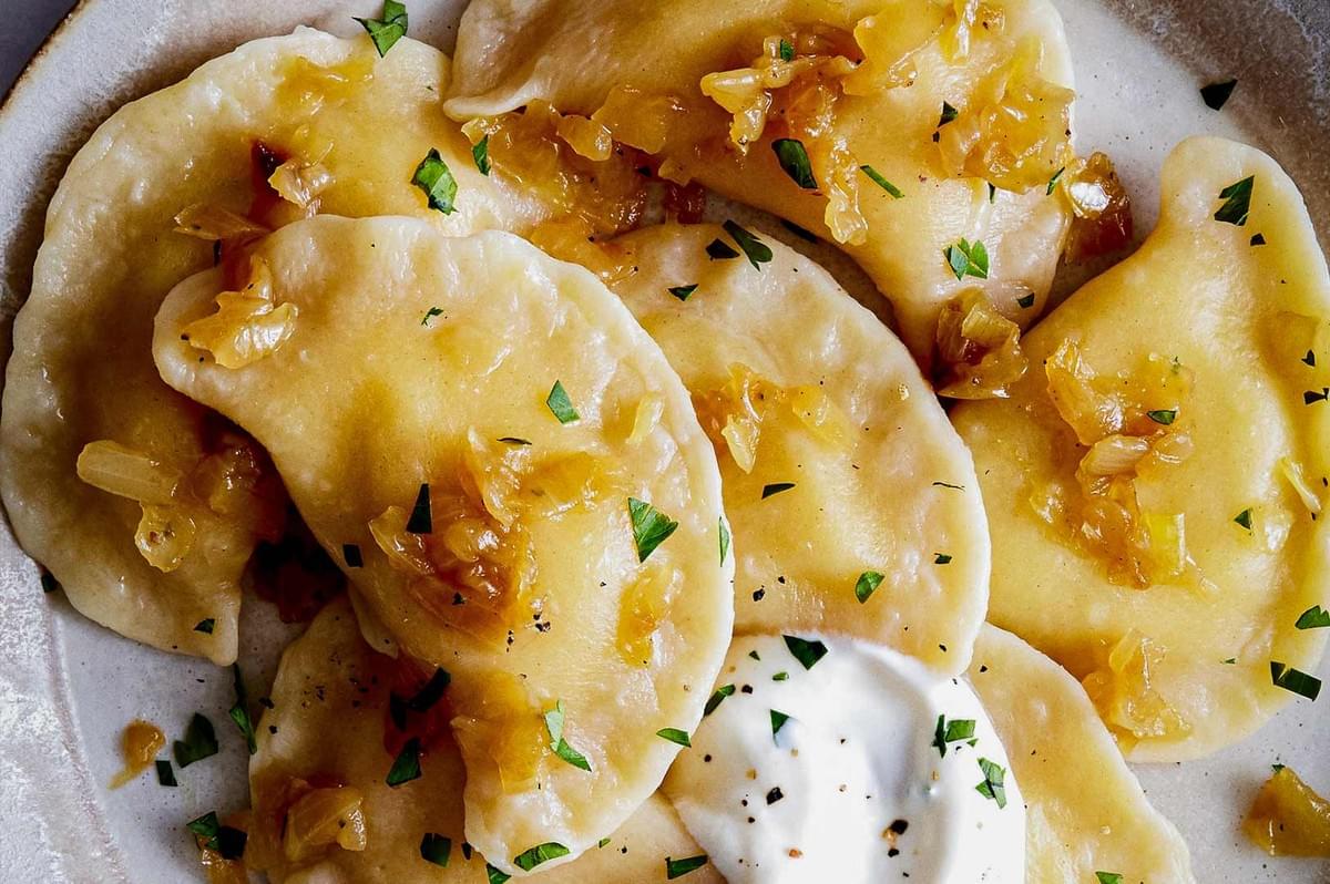 Polish pierogi dumplings topped with sautéed onions and parsley, served with sour cream on ceramic plate.