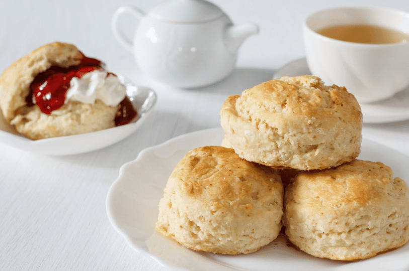 A stack of three plain scones on a white plate, positioned next to a teapot and a cup of tea, with one scone in the background already topped with cream and jam.