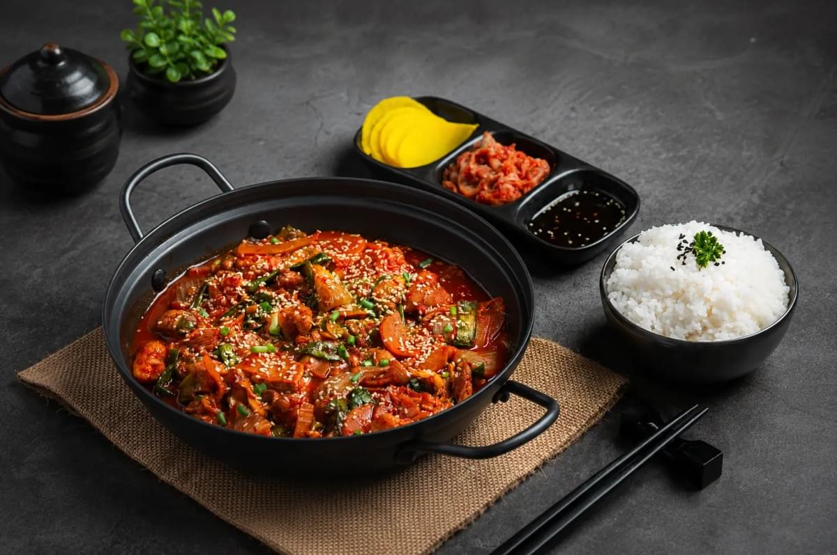 A black pan filled with a spicy, red Korean stir-fry garnished with sesame seeds, served alongside a bowl of white rice and small side dishes on a dark surface.