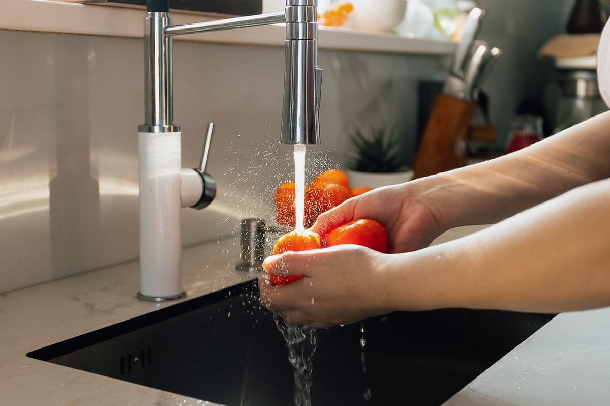 Close-up front angle shot of fresh vegetables being rinsed under running water in a kitchen sink, highlighting food safety practices and proper washing methods before food preparation and storage.