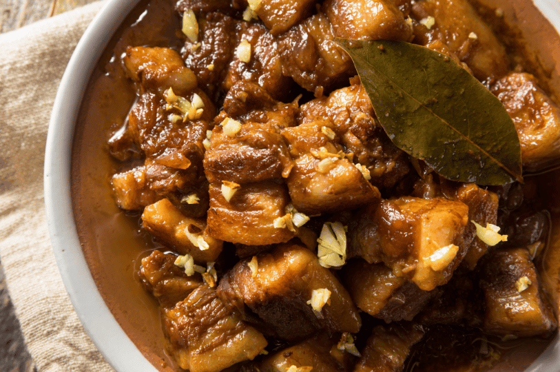 A close-up shot of Pork Adobo served in a shallow clay-colored bowl, generously garnished with toasted minced garlic and a bay leaf.