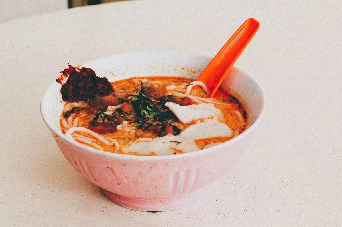 A bowl of Laksa soup with a spoon resting beside it, showcasing its rich, aromatic broth and garnishes.