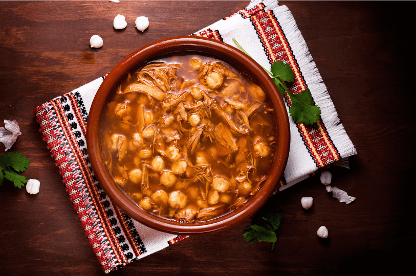 A warm, close-up shot of a brown ceramic bowl filled with hearty Pozole Rojo, showcasing the rich broth and plenty of softened hominy kernels.