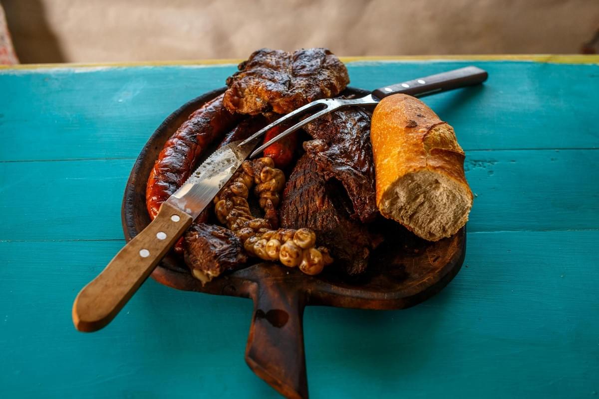  A wooden carving board sits on a vibrant teal surface, piled high with an assortment of grilled meats including sausages, ribs, and chitterlings. A large carving knife and fork are resting across the food next to a crusty piece of bread, ready for serving.