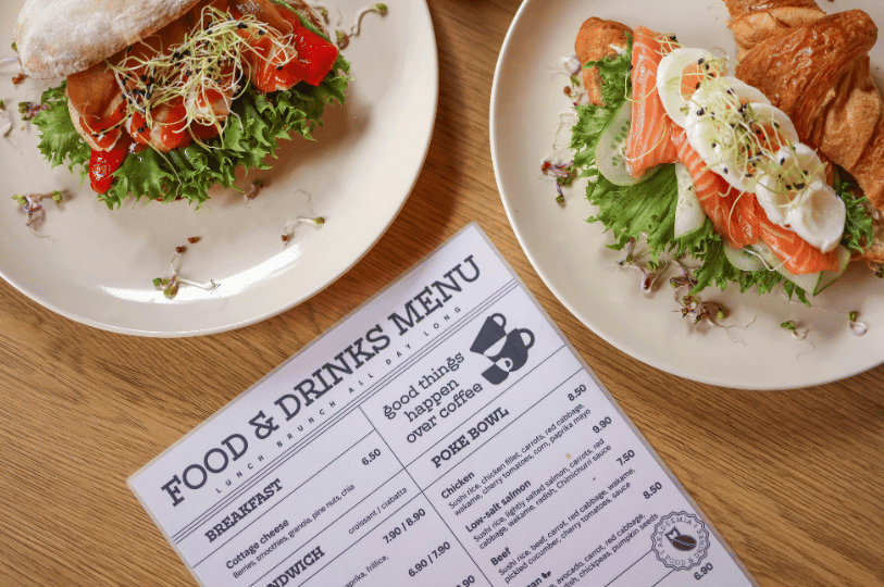 A casual "Food & Drinks Menu" resting on a wooden cafe table alongside two white plates—one holding a chicken and red pepper sandwich, and the other a smoked salmon and boiled egg croissant, both garnished with fresh microgreens.