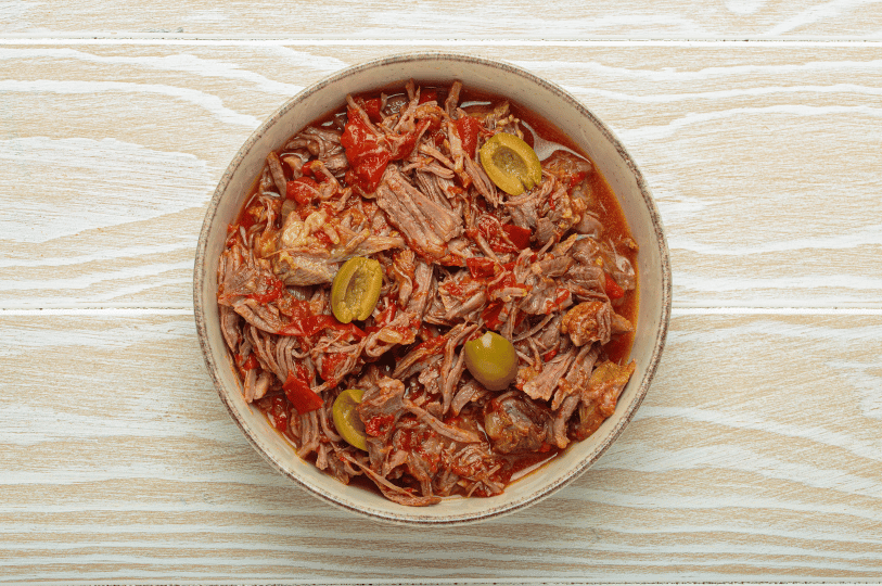A top-down shot of a ceramic bowl filled with traditional shredded Ropa Vieja beef, featuring visible strips of red pepper and topped with several green olives.