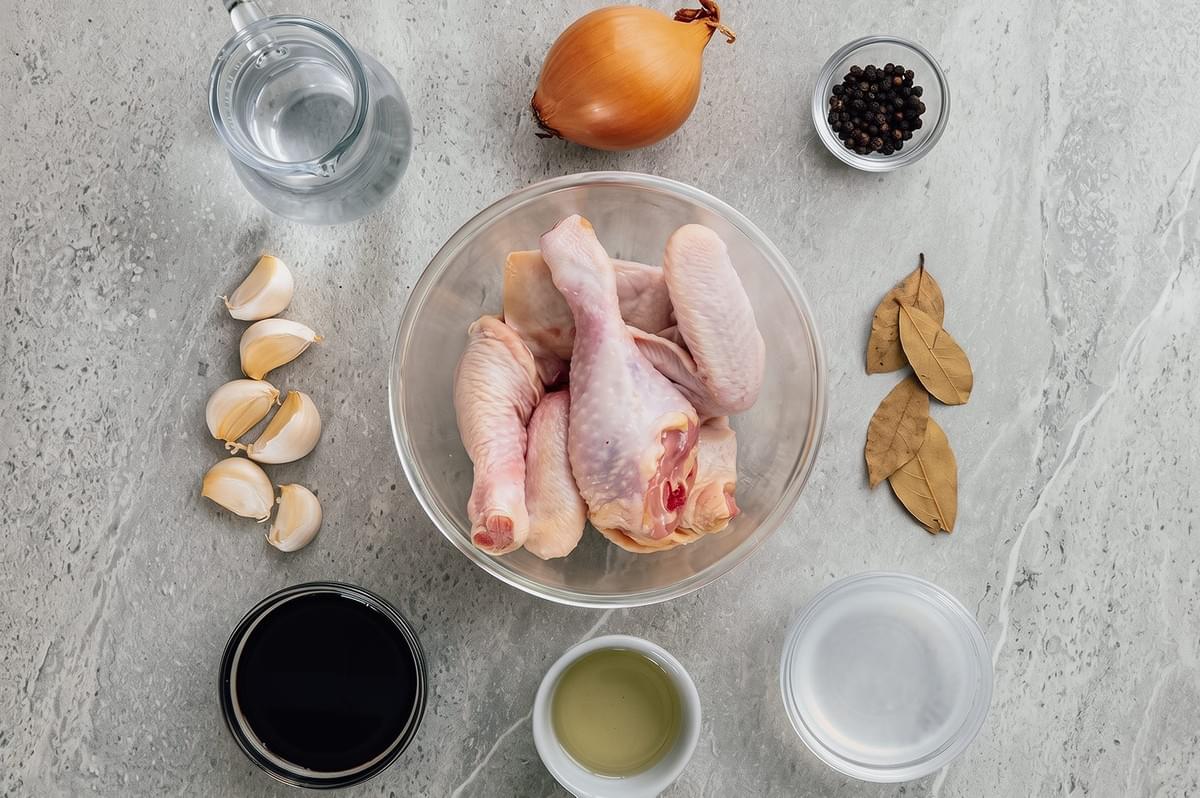Overhead flat lay of Filipino adobo ingredients including raw chicken, soy sauce, vinegar, garlic, onion, bay leaves, peppercorns, and water arranged for traditional chicken adobo preparation.