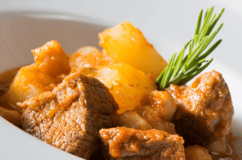 A close-up of chunky beef and potatoes simmered in a rich, reddish-orange sauce, garnished with a fresh sprig of rosemary in a white ceramic bowl.