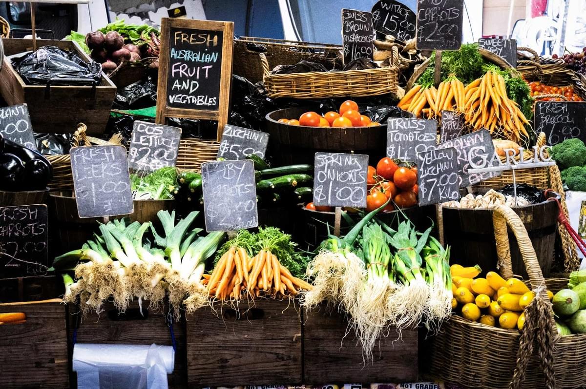 A farmers’ market stall overflowing with fresh produce including carrots, tomatoes, zucchinis, leeks, eggplants, and citrus fruits, with chalkboard signs showing prices.