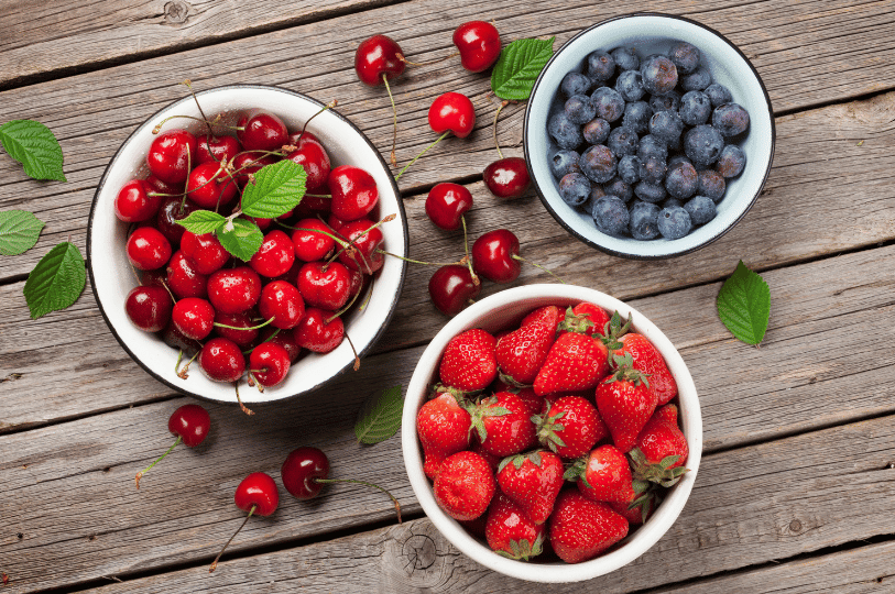 Three white enamel bowls on a weathered wooden table containing sorted cherries, blueberries, and strawberries, representing the organized mise en place needed before starting the canning process.