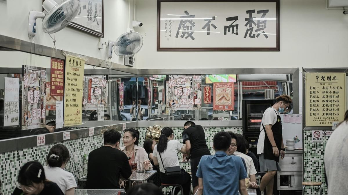 Inside a bustling restaurant characterized by green mosaic tiles and mirrored walls, numerous diners are gathered at small tables to enjoy their meals. A large framed piece of Chinese calligraphy hangs prominently above the scene, overlooking the lively atmosphere where customers chat and a server in an apron attends to the floor.
