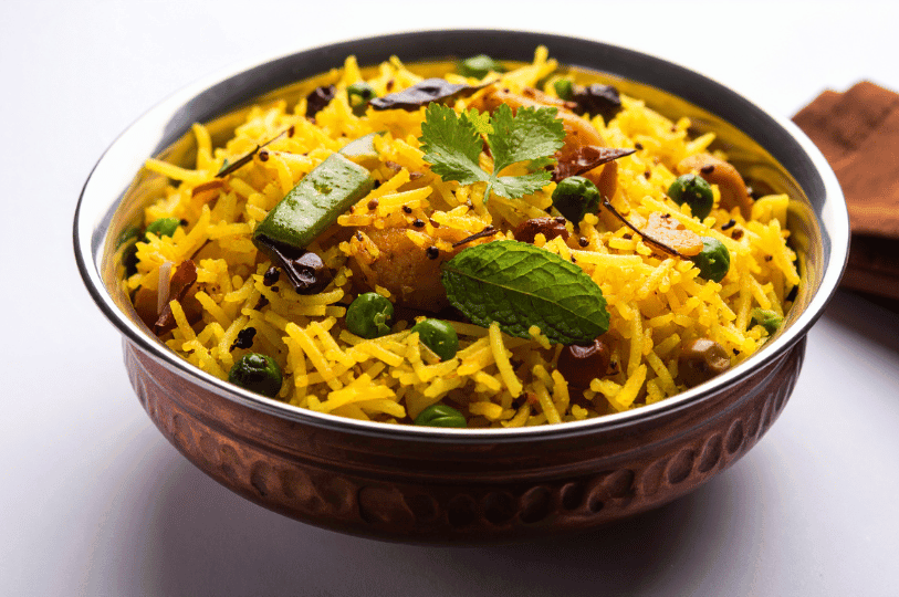 A metallic bowl filled with richly spiced yellow rice, generously mixed with green peas, dark spices, and topped with fresh herbs including mint and cilantro.