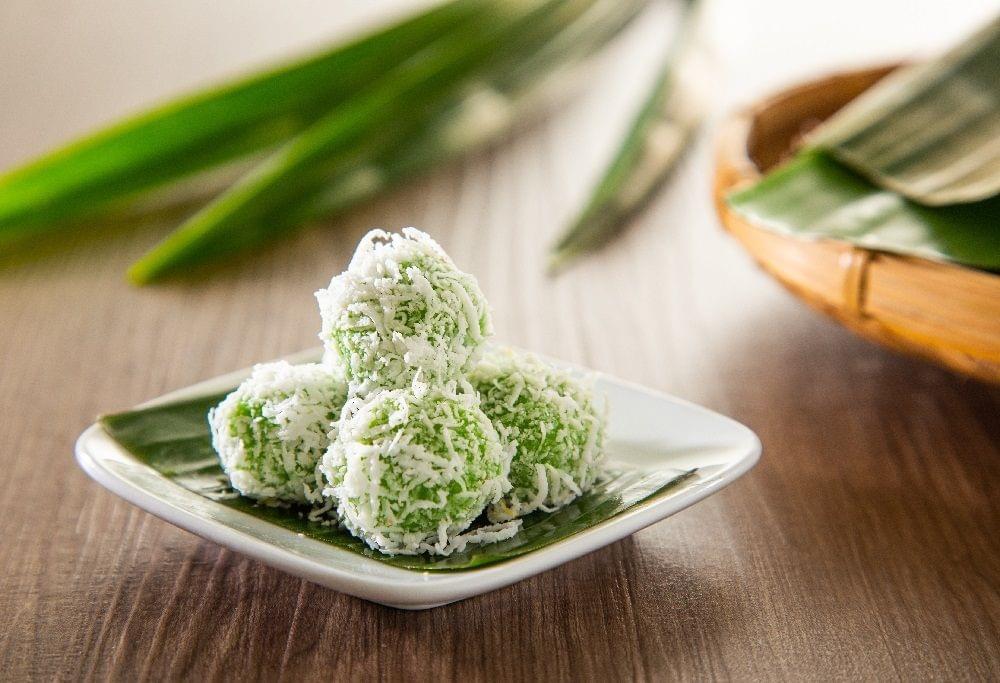 A stack of green, coconut-dusted ondeh-ondeh sits on a small white plate lined with a banana leaf. Long pandan leaves and a woven basket rest in the soft-focus background on a wooden tabletop.