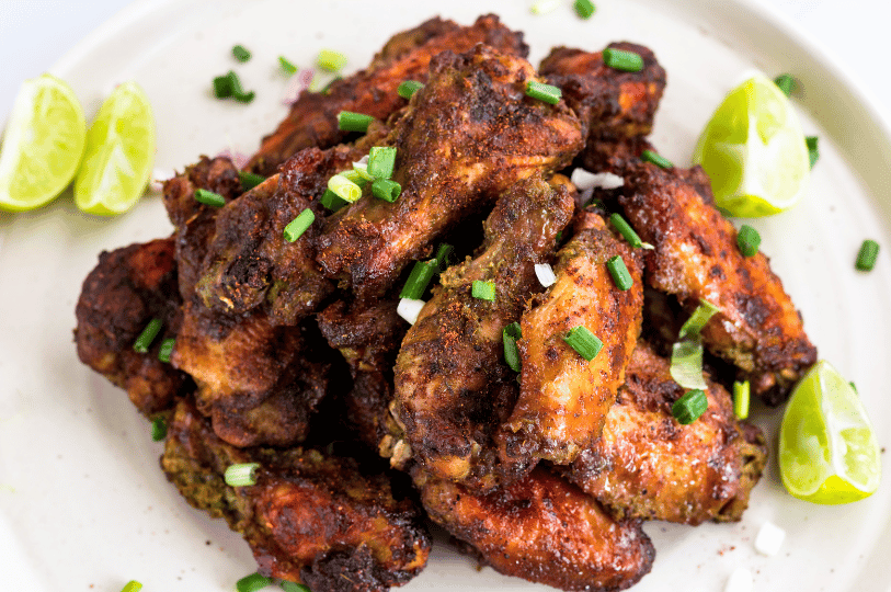 A close-up, high-angle shot of a pile of jerk chicken wings and drumettes seasoned with a dark spice rub and garnished with fresh scallions and lime wedges on a white platter.