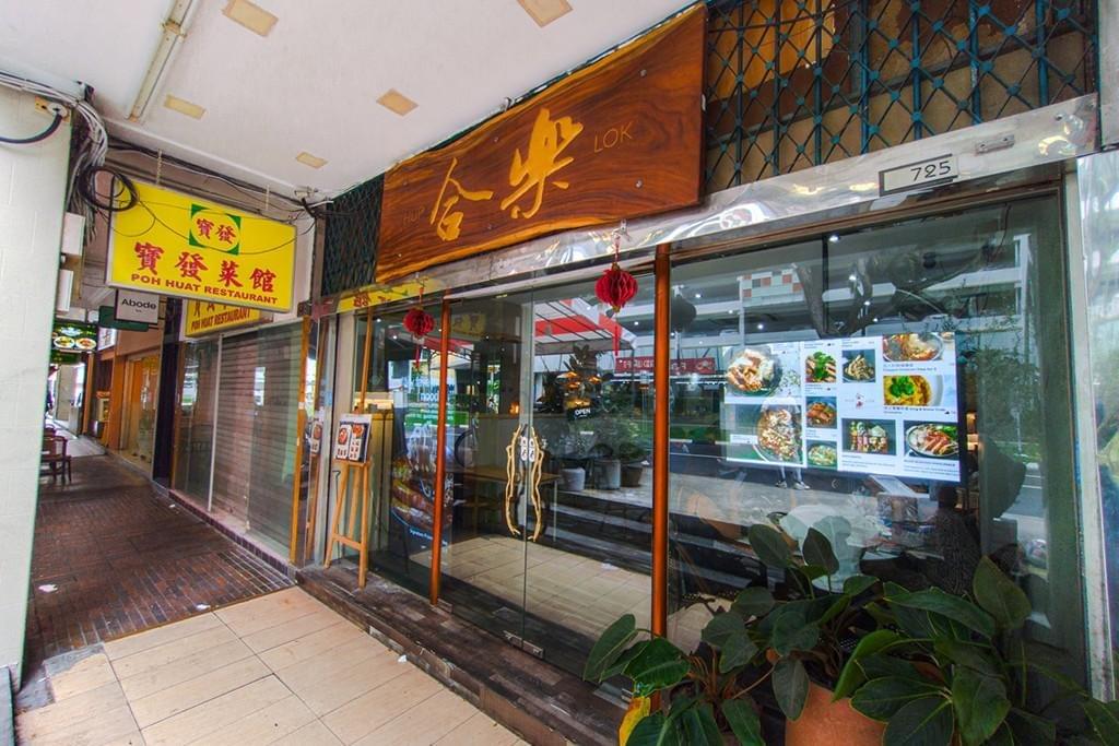 The storefront of Hup Lok restaurant features glass doors, a large wooden sign with gold Chinese characters, and a digital menu displayed in the window. Located along a covered walkway, it sits next to neighboring shops with brightly colored signs like Poh Huat Restaurant.