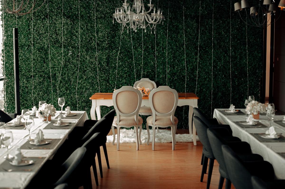 Elegantly decorated dining area in a halal restaurant featuring long tables with white tablecloths, modern dark chairs, and a romantic sweetheart table against a green plant wall with fairy lights.