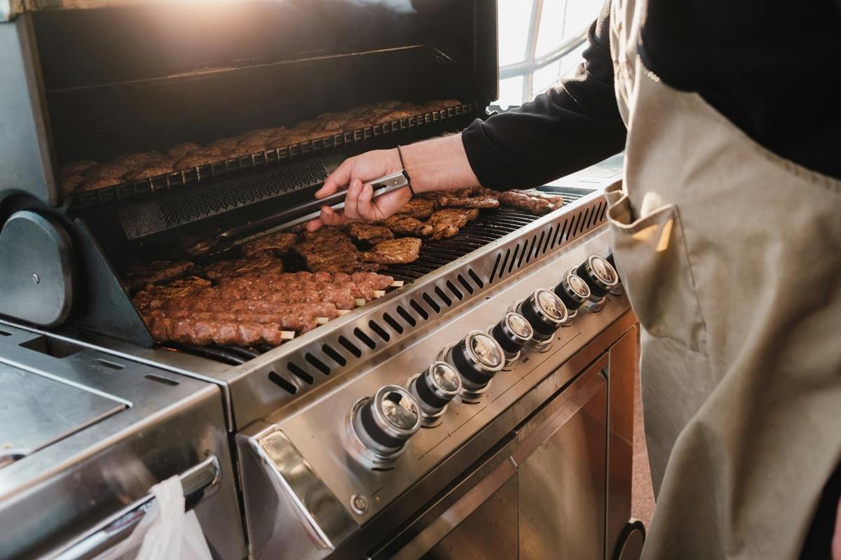  A person wearing a tan apron uses metal tongs to flip various meats, including patties and kebabs, on a large stainless steel outdoor grill. The scene is brightly lit by natural sunlight, highlighting the steam rising from the cooking food and the rows of control knobs on the front of the appliance.