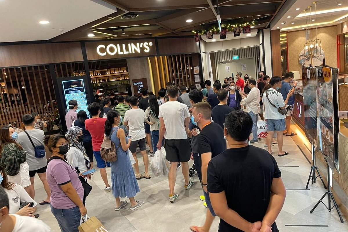 A large crowd of people forms a long, dense queue waiting in front of a restaurant named "COLLIN'S" inside a brightly lit shopping mall. Most people in the line are wearing casual clothing and face masks while facing away from the camera toward the restaurant entrance.