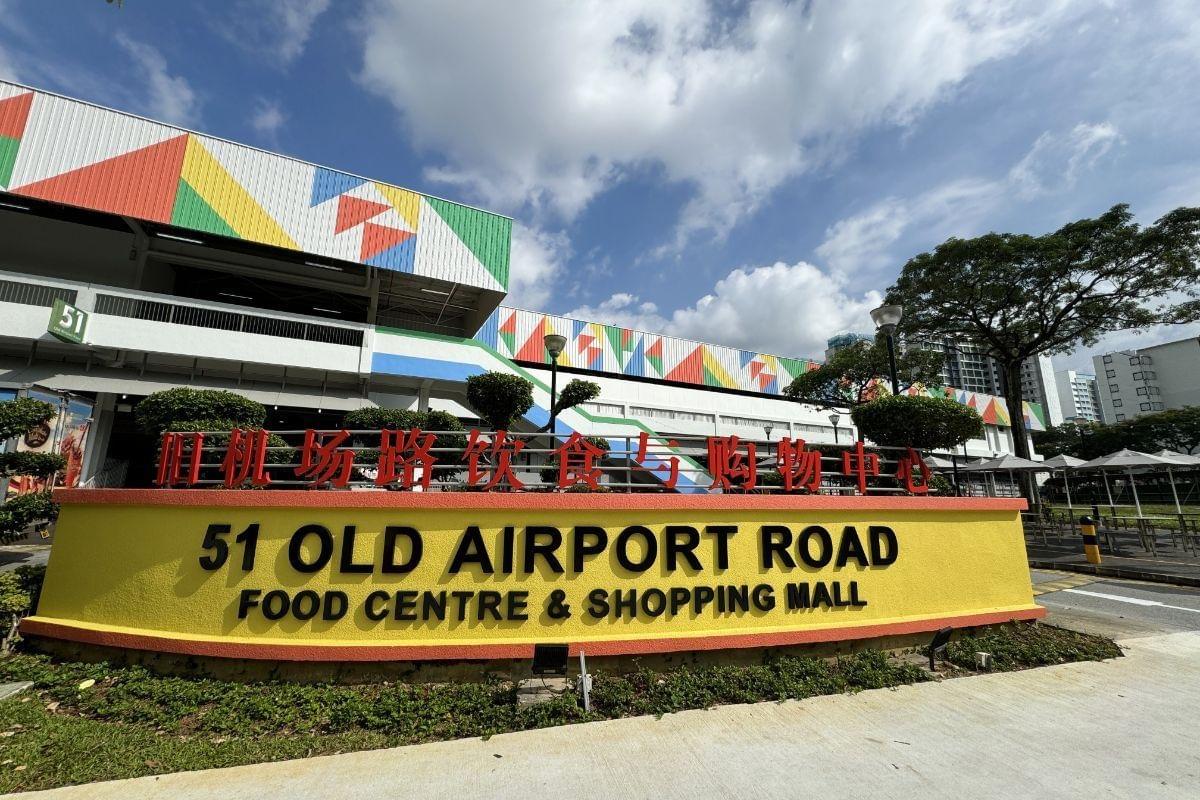 A large yellow sign identifies the entrance to the Old Airport Road Food Centre & Shopping Mall under a bright, cloudy sky. The building in the background features a modern facade decorated with colorful geometric patterns in red, green, and blue.