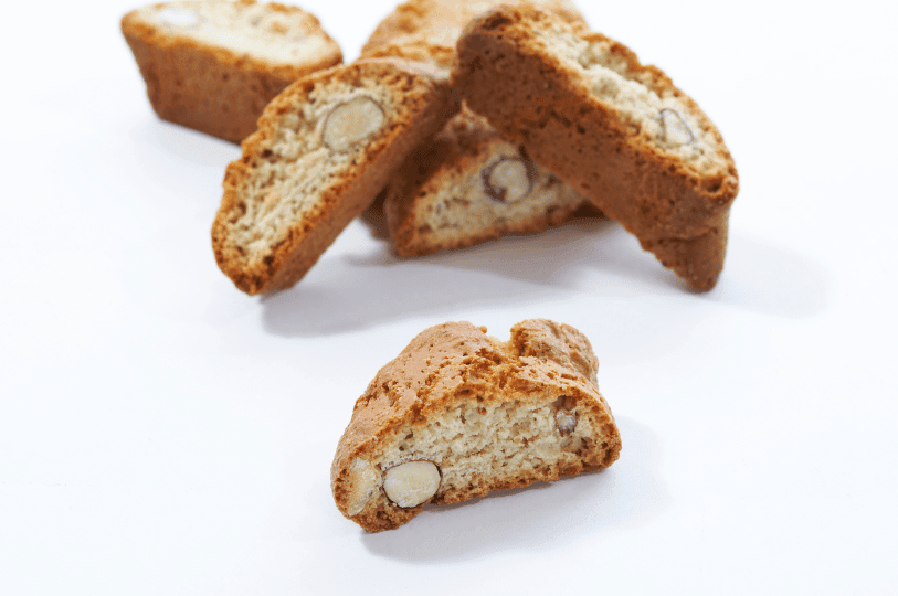 A crisp almond biscotti rests in sharp focus on a white surface, its golden hue and embedded nuts highlighting the twice-baked texture, while blurred pieces in the background evoke a cozy, coffee-paired indulgence.