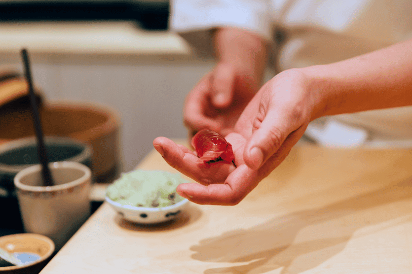 A sushi chef in a white uniform delicately places raw tuna atop vinegared rice at a wooden counter, surrounded by bowls of wasabi and soy sauce—capturing the precision and seasonal artistry of Japanese cuisine in Singapore.