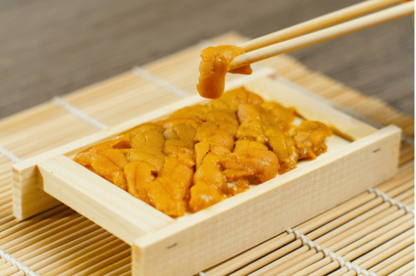 Chopsticks holding a delicate piece of sea urchin over a small wooden box filled with fresh uni, resting on a bamboo mat.