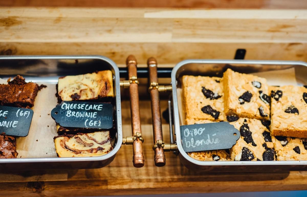 Rectangular metal bins present a selection of baked dessert bars, featuring swirled cheesecake brownies on the left and cookie-studded Oreo blondies on the right. Rustic black tags label the items, indicating the brownies are gluten-free, while copper pipe handles separate the trays on a light wooden surface.