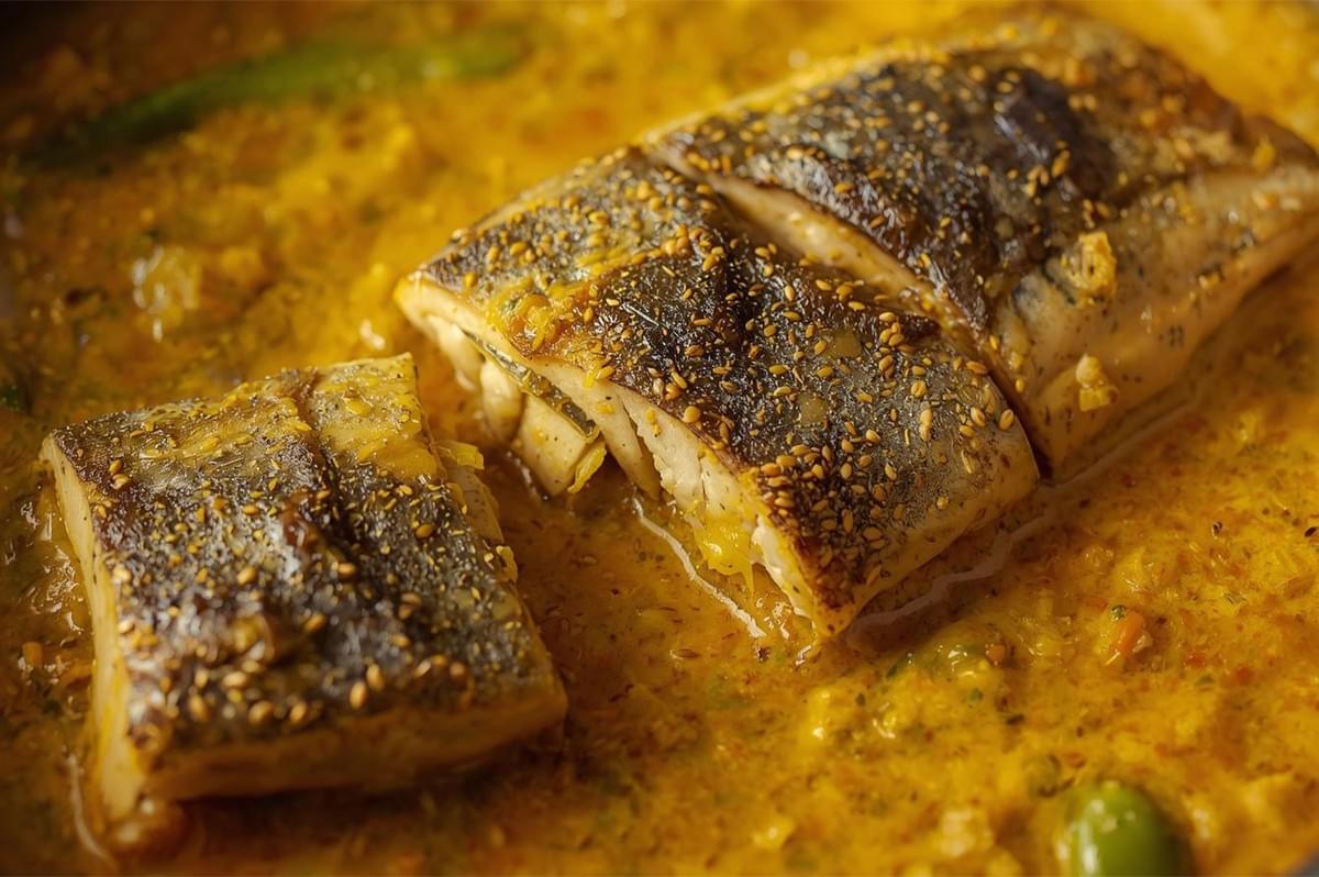 A close-up of a long piece of firm white fish coated heavily in black and white mustard seeds and cooked in a vibrant yellow-orange, oily sauce (likely Shorshe Maach or a Bengali-style mustard fish curry).