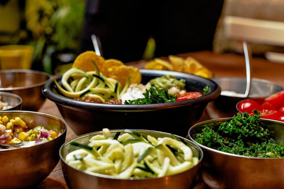A large dark bowl is filled with vibrant ingredients like zucchini noodles, kale, guacamole, and tortilla chips. Smaller metal bowls containing fresh corn salsa, sliced tomatoes, and additional greens surround the main dish on a warm wooden table.