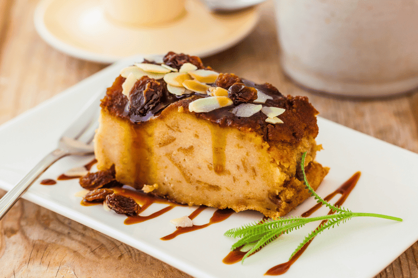 A gourmet slice of bread pudding on a white plate, drizzled with caramel sauce and topped with raisins and toasted almond slivers.