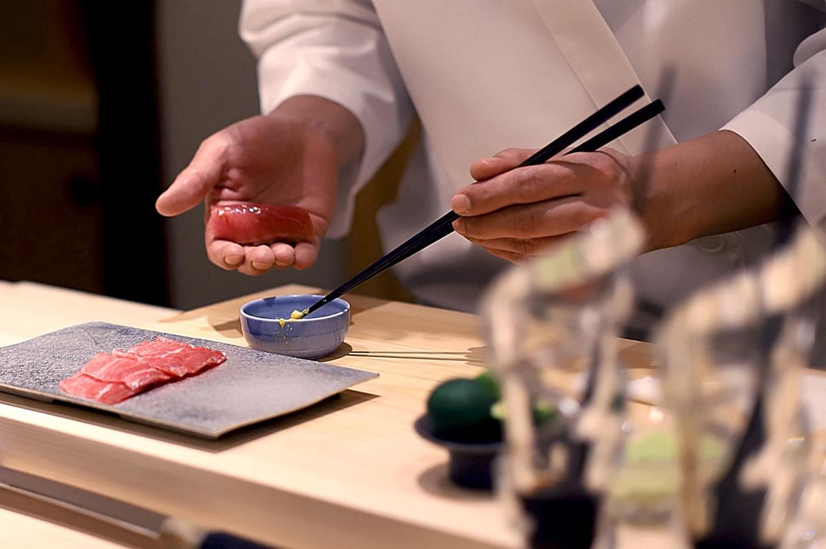 Chef in a white coat expertly prepares sushi, using chopsticks to add wasabi. Fresh slices of tuna rest on a gray plate, evoking precision and care.