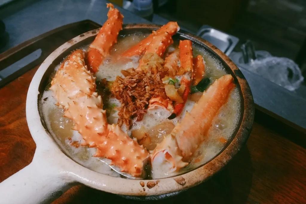 A steaming clay pot filled with hot broth and large, spiky orange king crab legs. The soup is garnished with fried shallots in the center, sitting on a dark wooden tabletop.