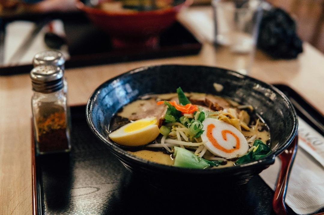 A steaming black bowl of ramen sits on a dark tray in the foreground, topped with a soft-boiled egg, narutomaki fish cake, chashu pork, and fresh greens. Small spice shakers sit to the left of the bowl on a wooden restaurant table, with another blurred bowl of ramen visible in the background.