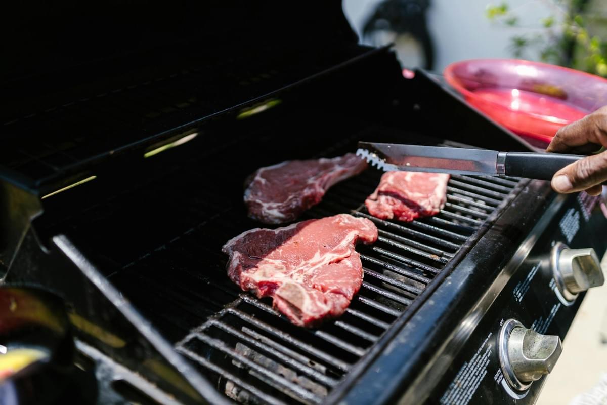 Three thick, raw steaks are being cooked on a black outdoor grill, with a person's hand using metal tongs to adjust one of the cuts. The grill features silver control knobs on the front panel and sits near a red bowl in a sunlit outdoor setting.